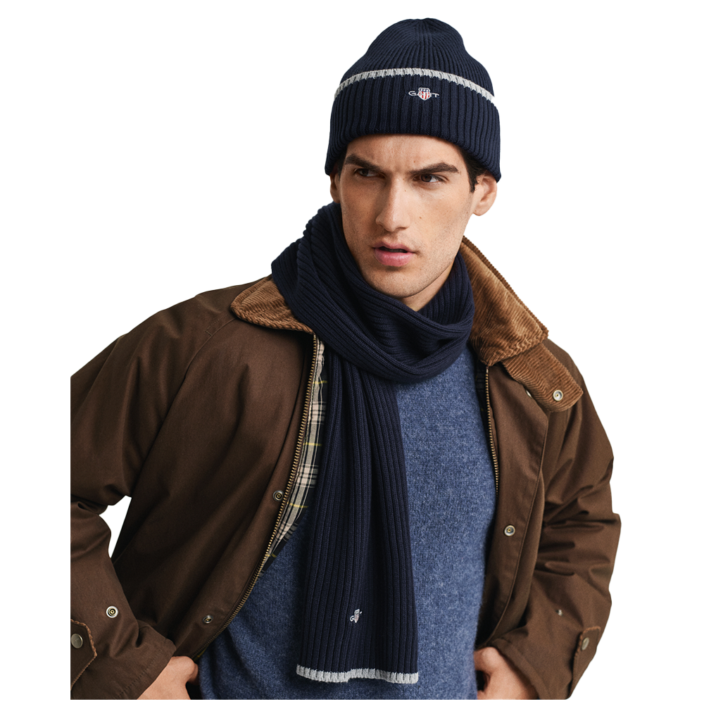 Man wearing a brown jacket, blue sweater, navy scarf, and beanie with a brand logo.