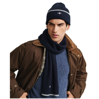 Man wearing a brown jacket, blue sweater, navy scarf, and beanie with a brand logo.