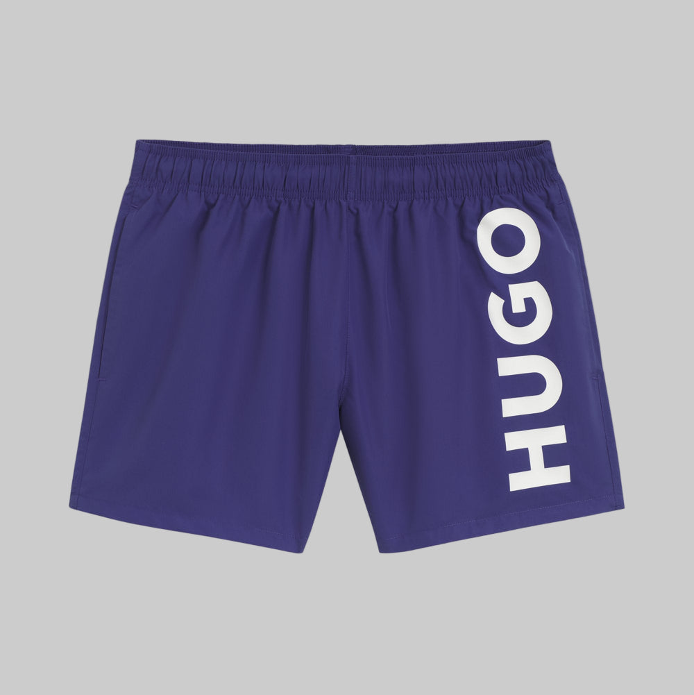 A pair of medium blue swim shorts with a white vertical logo print on the right leg, branded with the name 'HUGO'.
